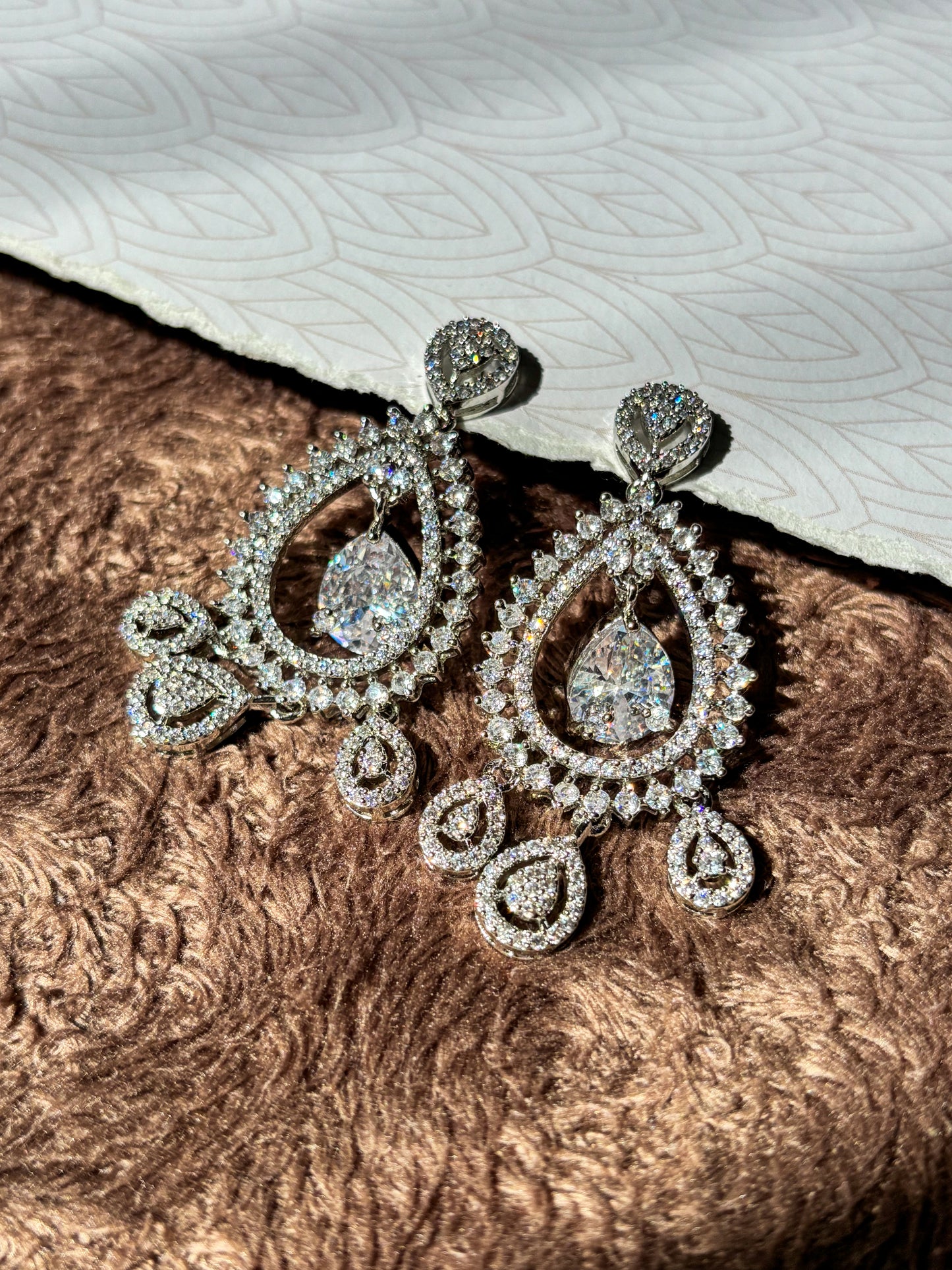 Sparkling silver-tone American Diamond (AD) Chandbali earrings featuring a large teardrop-cut crystal centerpiece, intricate pavé stone detailing and elegant dangling accents. Perfect for bridal wear, weddings, festive occasions and party styling.