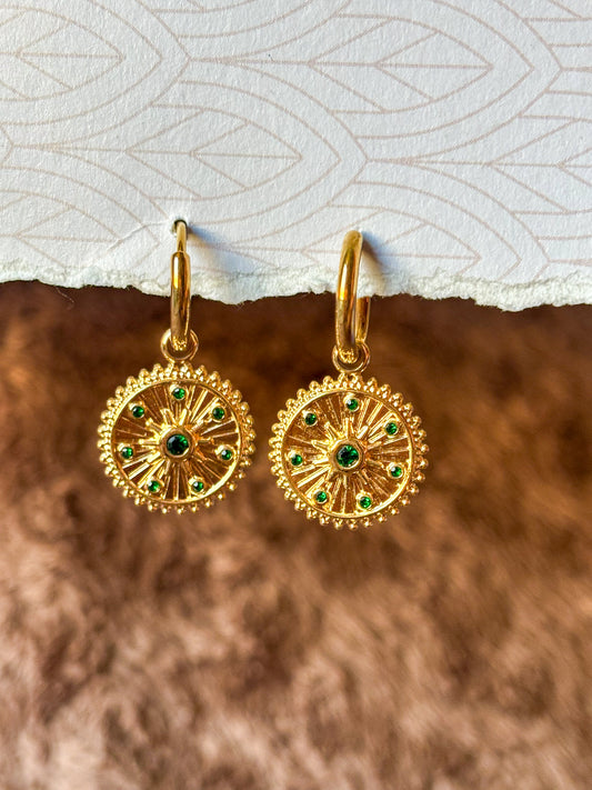 Close-up of gold-tone sunburst hoop earrings featuring intricate radial detailing and sparkling green gemstone accents. Elegant vintage-inspired dangle earrings
