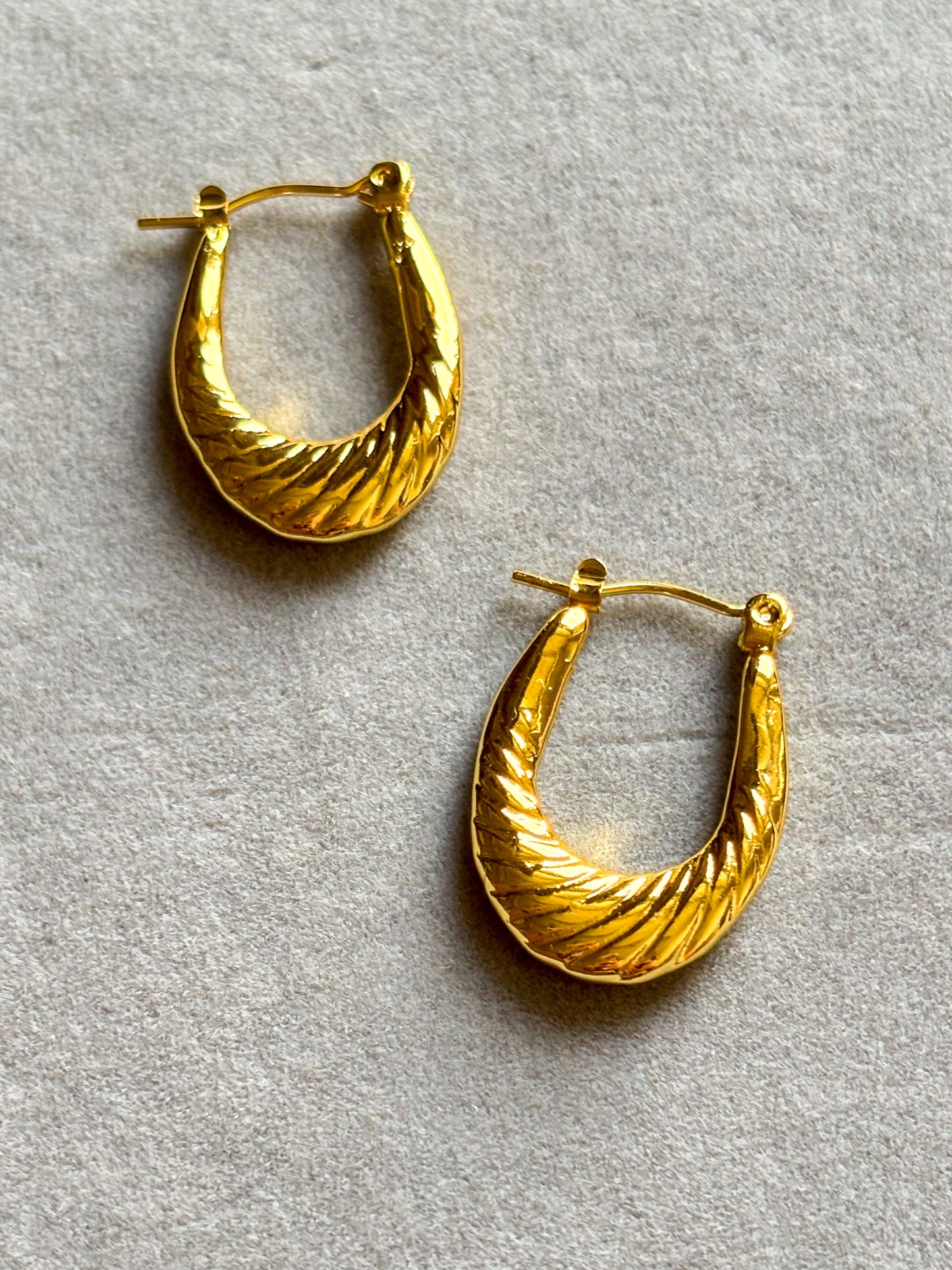 Anti tarnish textured gold hoop earrings with a sculpted twisted design, displayed on a soft neutral aesthetic background. Lightweight waterproof statement hoops with a polished gold finish for everyday wear.