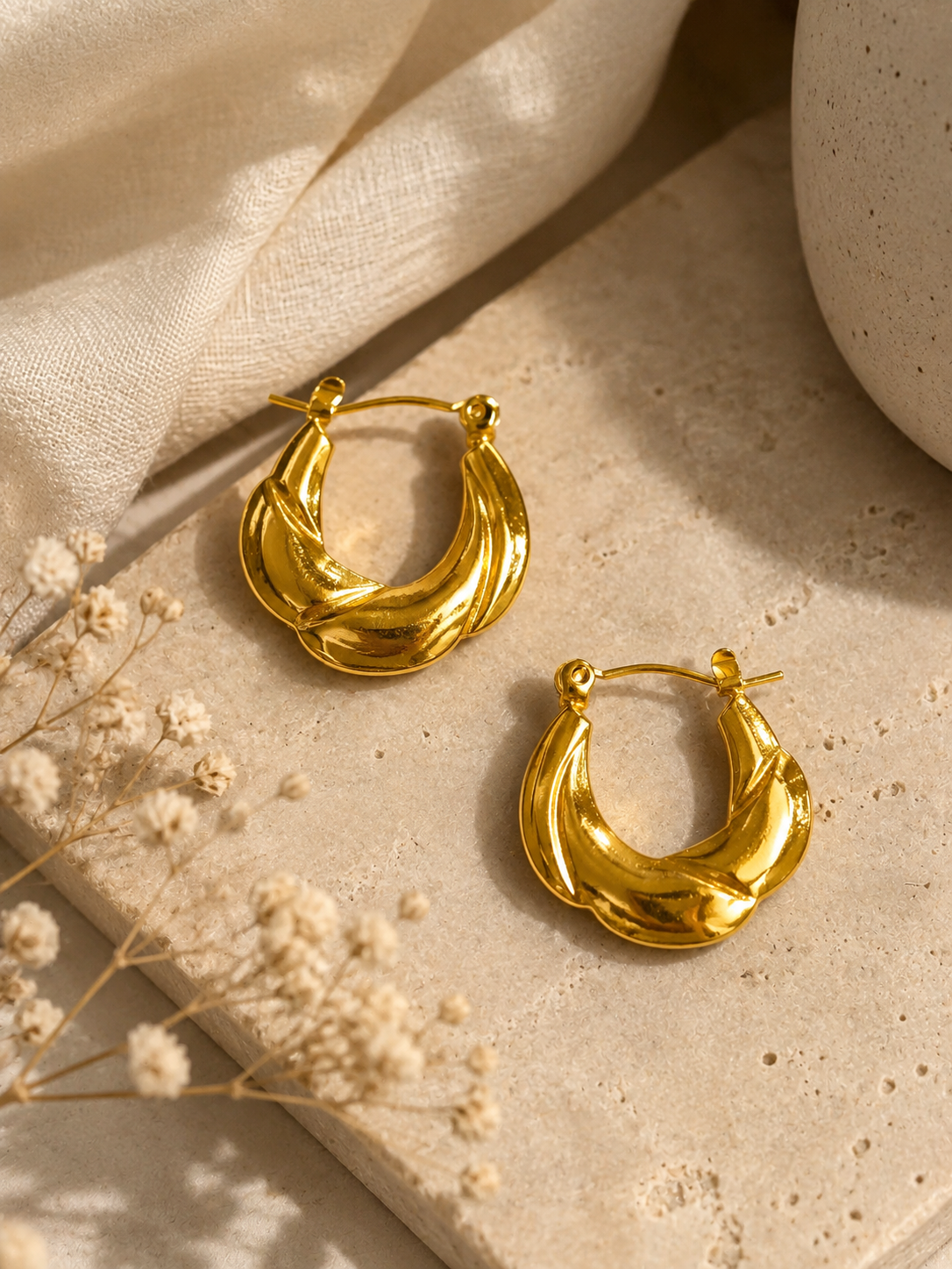 Anti-tarnish sculpted gold hoop earrings with a bold textured design, displayed on a warm beige aesthetic stone background with soft fabric, dried flowers and natural lighting for a luxurious, minimalist look.
