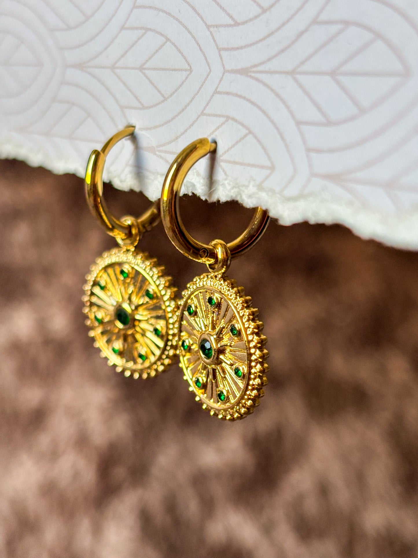 Close-up of gold-tone sunburst hoop earrings featuring intricate radial detailing and sparkling green gemstone accents. Elegant vintage-inspired dangle earrings
