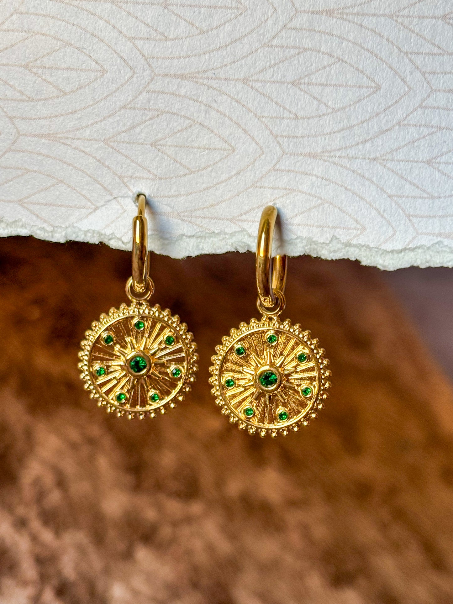 Close-up of gold-tone sunburst hoop earrings featuring intricate radial detailing and sparkling green gemstone accents. Elegant vintage-inspired dangle earrings